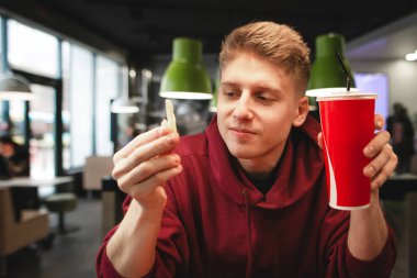 Çekici bir genç adam elinde, bir bardak patates ile bir fast-food restoran oturur yemek görünüyor ve gülümseyerek. Fransız kızartması yemek mutlu bir genç adam portresi.