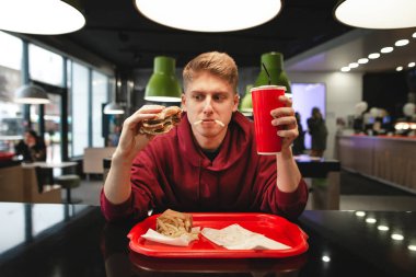 Bir komik genç adam yeme fast food restoranın arka plan, portresi. Komik aç öğrenci bir aperatif yiyecek ile bir restoranda hamburger ve patates kızartması yiyor.