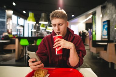 Adam bir içecek kırmızı cam içecekler ve bir smartphone bir lokanta kafede kullanır. Bir akıllı telefon istimal belgili tanımlık enterne ve içme çekici bir çocuk onun elinde büyük bir kırmızı cam ile portresi
