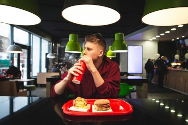 Bir fast-food restoranında elinde bir tepsi yiyecekle oturmuş kırmızı bardaktan soğuk bir içecek içen ve gözlerini kaçıran genç bir adamın portresi. Restoranda öğrenci yemekleri..