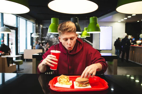 一个有趣的年轻人坐在快餐店里 拿着一杯饮料 从托盘里拿了一个汉堡的肖像 青少年在快餐店吃美味有害的食物 — 图库照片