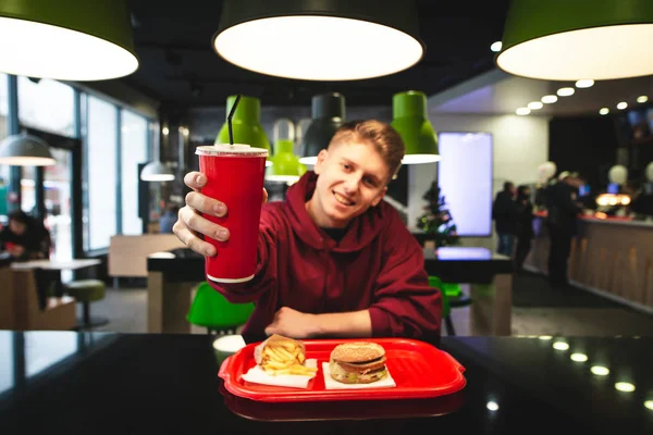 Smiling young man sits at the table with a fast food tray, holds a glass of drink in his hand and shows in the camera. Happy student eats fast food, shows a glass. Focus on the red glass in his hands.