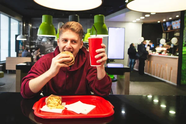 Positive teen in street clothes is eating fast food at a fast-food restaurant, biting a burger, looking at a glass of drink and smiling. Funny student with a fast food tray eating at the restaurant