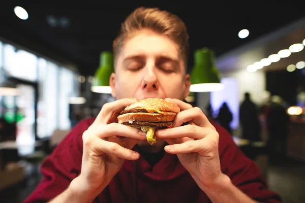 年轻人吃了一个美味的大汉堡 他的眼睛闭着 一个有趣的男人咬汉堡的特写镜头 快餐店的有害美味食品 — 图库照片
