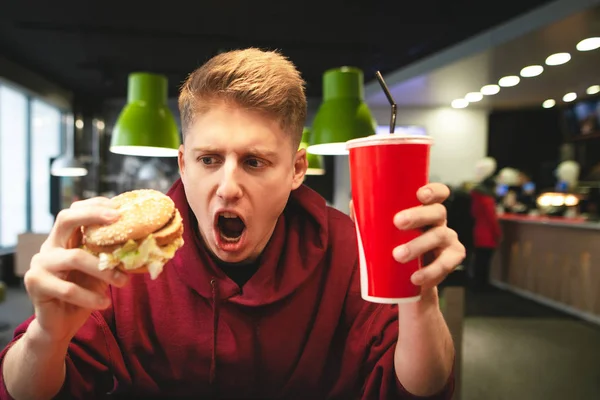 Joven Emocional Con Vaso Bebida Una Hamburguesa Mano Mira Hamburguesa — Foto de Stock