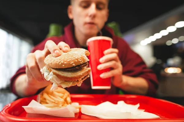 Genç adam, bir fast food restoran yemekleri lokanta. Adam bir burger kırmızı cam dışında bir kola içme, elinde tutar. Patates kızartması ve lokanta tepsi. Fast Food kavramı. Genç fast food yiyor
