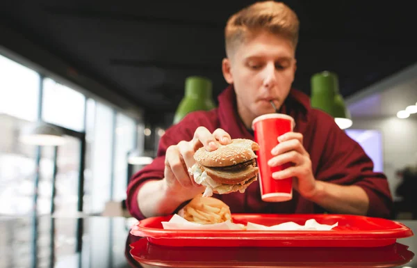 Tray with fast food, french fries, burger in the hands of a young man drinking a drink from a red glass. Fast Food Concept. Student dishes fast food at the fast food restaurant.