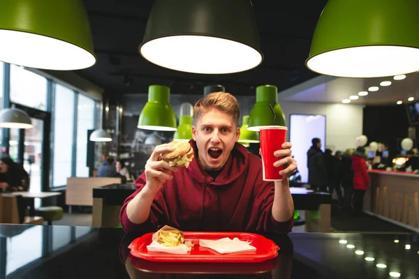 Emotional student sitting at the fast food restaurant table, holding an appetizing burger and a glass of glass in his hands, looks at the camera with astonished look. Harmful food and young man.
