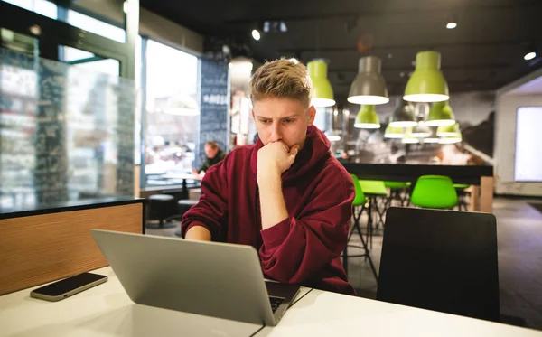 Young business man in casual clothing works in a cafe on a laptop, focuses on looking at the screen. Freelancer works in a fast food cafe.