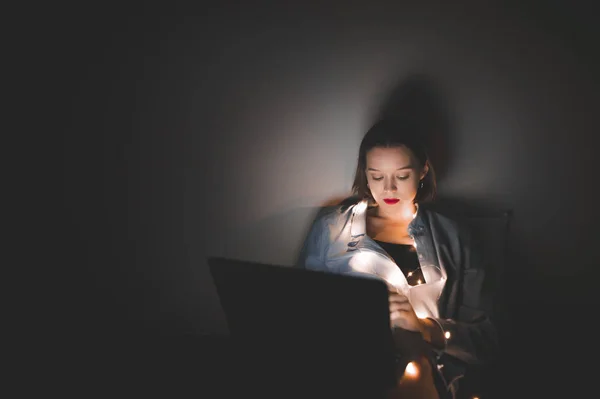 Woman with a laptop on her legs, lying in bed at Christmas lights at night, uses a laptop. Focus is on the girl hands works on a laptop at night. Gadget and technology concept. Night work