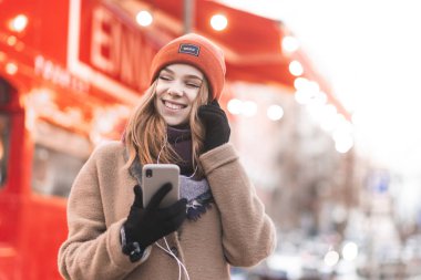 Mutlu kız ceket sokakta yürüyor, müzik kulaklık kapalı gözleriyle dinler ve smartphone elinde tutar. Güzel kız sıcak giysiler ve kulaklık güzel bir arka plan üzerinde