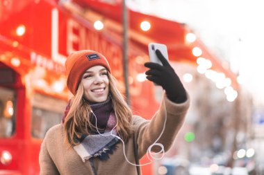 Mutlu kadın yakınındaki bir smartphone cep telefonu ile kırmızı bir otobüs sokak gülümsüyor ve alır selfie arka planda duran sıcak giysiler içinde. Güzel pozitif kız selfie bir kış yürüyüş alır.