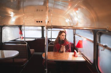 Güzel bir kadın ile özgün bir retro iç bir kafede oturan bir battaniye sarılı. Kız kafe otobüs oturuyor, bir fincan kahve elleri ve warmes, Vitrinlere bakıyor tutar