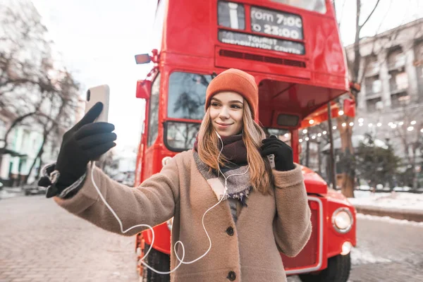 Girl in a coat and headphones standing outdoors against a background of red bus and take selfie. Happy woman is a tourist in warm clothes, photographed on the background of a red bus.