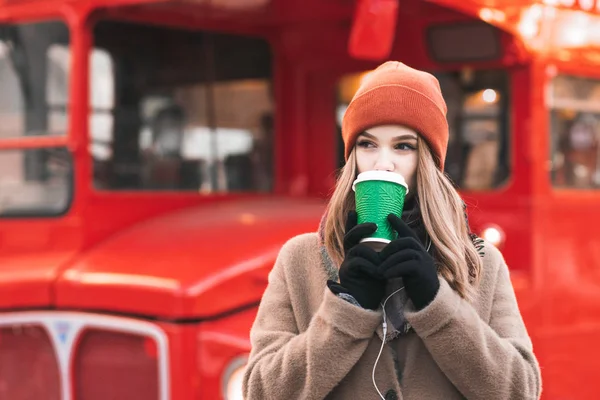 暖かいコートとオレンジの帽子 赤バスの背景の通りに立って 紙コップのコーヒーを飲んで 横て女性を挙げます Copyspace コーヒーを飲みながら甘い学生の女の子 — ストック写真