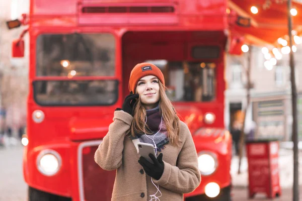 Cute girl coat and hats stand on the background of the red bus,listen music in the headphones, hold the smartphone in hand and look up.Young beautiful woman in warm clothes on a red street background.