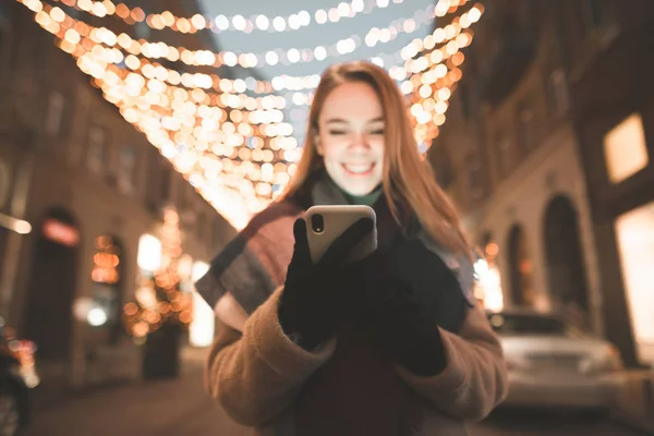 Night portrait of a cute girl holds a smartphone in her hands and looks at the screen , backdrop of city lights decorations. Happy woman uses phone at night on the street.Focus on the smartphone