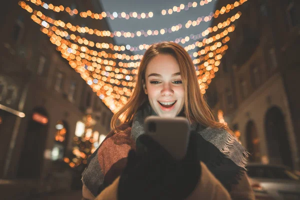 Close-up portrait of a surprised girl looks at the smartphone screen and smiles, background of the night street lights. Attractive woman emotionally looks at the smartphone,background night landscape