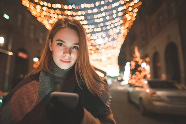 暖かい服で美しい女性は カメラに探してナイト ストリートの背景に彼女の手でスマート フォンと立っています 街路灯装飾の背景の彼女の手で携帯電話を持つ女性 — ストック写真