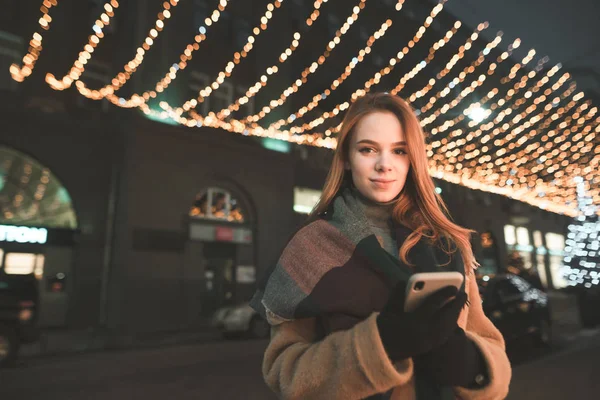 コートで美しい少女は 街路灯の飾りの背景にスマート フォンを使用します 暖かい服で女性の肖像画はスマート フォンを使用して 画面と笑顔で夜の散歩では — ストック写真