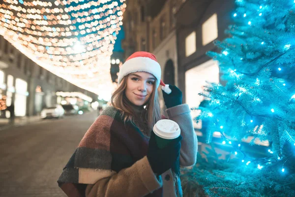 Smiling sweet girl in a Christmas hat and a cup of coffee in her hands stands at the Christmas tree on the street, looks at the camera and smiles. Christmas concept.