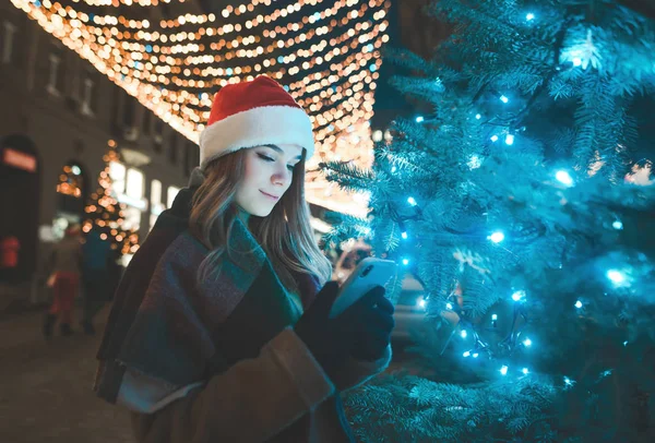 Sweet woman in a Christmas hat stands on a tree on a street decorated on a holiday and uses a smartphone. Christmas portrait of a beautiful girl using a smartphone near a Christmas tree , street