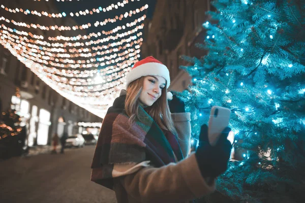 Beautiful girl in a Christmas hat stands at night on a street near a Christmas tree and take selfie.New Year\'s portrait of a girl on a decorated street makes a photo on a smartphone.Christmas selfie