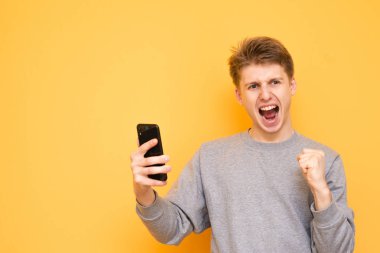 Sarı bir arka plan üzerinde duran ve kazanan bir jest yapma ve kameraya bakarak duygusal bir adam portresi. Adam sarı bir arka plan üzerinde izole onun elinde bir smartphone için bayram yapıyor