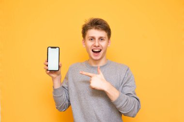 Mutlu adam bir akıllı telefon elinde tutan, parmağını gülümsüyor beyaz ekranda gösterir ve kameraya benziyor. Modern bir akıllı telefon ile neşeli adam sarı bir arka plan üzerinde izole.
