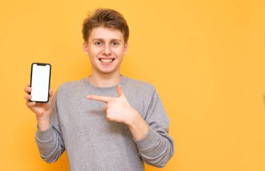 Mutlu adam elinde, modern bir akıllı telefon ile izole kameraya bakıyor. Gülümseyen adam telefonu elinde tutar, bir parmak bir smartphone ile sarı bir arka plan üzerinde beyaz bir ekran gösterir