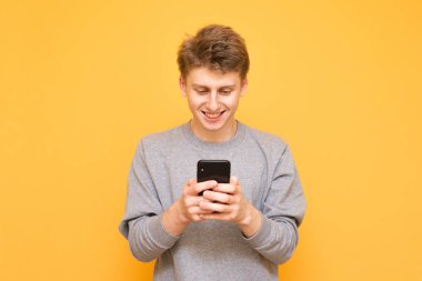 Sarı bir arka plan üzerinde duran, bir smartphone kullanarak ve ekrana bakarak mutlu bir adam portresi. Genç adam gülümseyen bir smartphone cep telefonu ile sarı bir arka plan üzerinde izole, Internet'i kullanır.