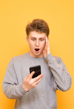 Şok adam bir smartphone üzerinde sarı bir arka plan kullanır, vasıl belgili tanımlık perde şaşkın bakışla görünüyor. Genç bir adam elinde bir akıllı telefon ile sürpriz kafasını ele aldı. Boşaltmak