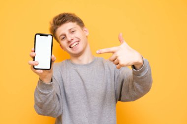 Mutlu çocuk sarı bir arka plan üzerinde duran, kameraya doğru görünüyor ve Smartphone parmak elinde tutan gösterir. Pozitif genç adam gülümseyen bir boş ekran smartphone gösterir. Boşaltmak