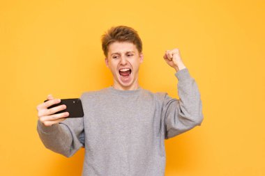Etkileyici adam oynarken bir smartphone oyun ve sarı bir arka plan üzerinde zafer sevinir. Mutlu genç adam bir akıllı telefon elinde tutar, vasıl belgili tanımlık perde görünüyor ve duygusal sevinir.