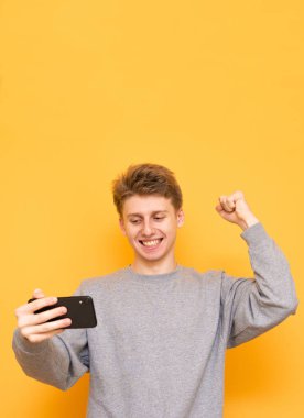 Mutlu bir adam bir akıllı telefon elinde tutar, ekrana bakıp duygusal sevinir, sarı bir arka plan üzerinde izole. Genç adam bir smartphone üzerinde sarı bir arka plan üzerinde Oyunlar zaferle sevinir