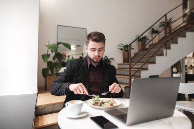 Bir restoranda iş man yemek kahvaltı. Tabak salata, bir dizüstü bilgisayar, bir fincan kahve, bir smartphone Restoran tablo. Adam bir çatal ve bıçak ile bir tabak Salata yiyor.