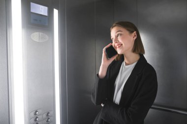 Genç gülümseyen işkadını yanında onun smartphone asansörde konuşuyor. Asansör telefonla bahsediyoruz işçisi kadın ofis.