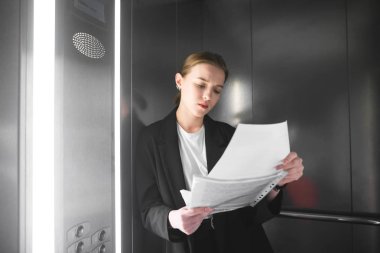 Kadın finansal analist eğitim belgeleri holding ayakta asansörde makaleler. İddialı işkadını Asansör belgelerinde dikkatle okuyor. Ciddi ve yoğun yüz.