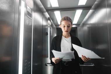 Genç bir kadın işçiye asansörde ayakta belgeleri okuma portresi. Konsantre işkadını Asansör belgelerinde denetliyor.