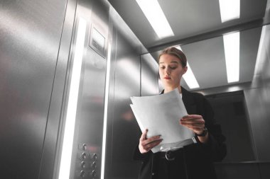 Meraklı kurumsal iş kadını kuşkuyla bakıyor belgeleri Asansör ayakta. Genç kadın işveren kritik Asansör gazetelerde okuyor. İnsan yüzü ifade, beden dili, tutum.