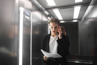 Rahat işkadını smartphone üzerinde konuşmak ve Asansör belgelerde okuma. Gazeteleri kontrol ve bir patronla telefonla konuşurken işçisi kadın ofis konsantre.