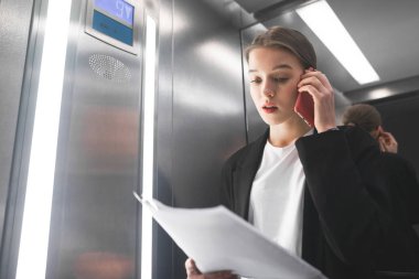 Asansörde belgeleri okuma smatphone ile rahat bir iş kadını portre kadar yakın. Evet ve asansörde belgelerine bakarak başarılı çalışan portresi.