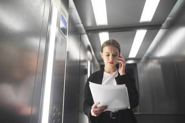 Ciddi iş kadını asansöre gazetelerde bakarak telefonla konuşuyor. Genç kadın işçiye belgeleri okuma ve smartphone Asansör tarafından konuşuyor.