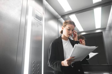 İşkadını konferans asansörde belgeleri okuma smartphone ile meşgul. Konsantre ofis çalışanı müşteriyle telefonla iletişim kurar ve gazeteleri okur. Arka plan.