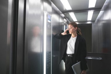 Memnun işkadını dizüstü elinde tutan asansörde onun Smartphone cep telefonu ile konuşuyor. Sinirli ofis çalışanı iş sorunları ve onun defter tutmak telefonla konuşuyor.