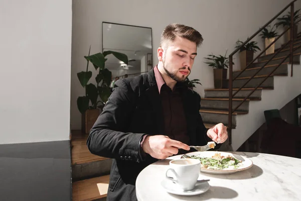 Man in a suit sitting at a table in a cozy restaurant with a cup of coffee and eating a delicious salad with a fork and a knife. Business man eats salad in a restaurant. Healthy Eating. Copyspace