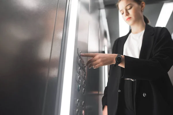 Diligent businesswoman pressing button inside elevator with a watch on her wrist. Female office worker in a black suit is pushing the button with her forefinger.