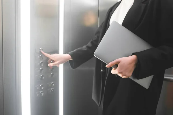 Female employee pressing the elevator button and holding the laptop. Businesswoman in the formal black suit is pushing the button of the lift and keeping her notebook.