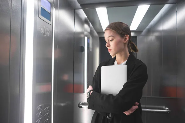 若い実業家は 彼女のラップトップを保持しているエレベーターの階数で Lookinig に集中されます エレベーターで女性事務員がよそ見彼女の手のノートを維持します — ストック写真