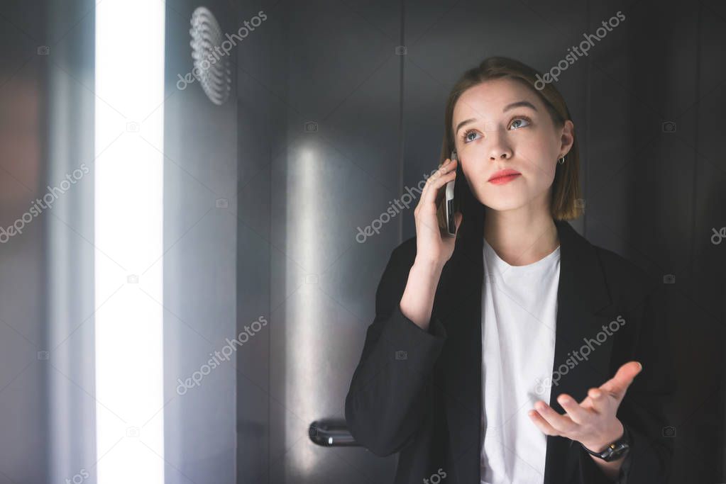 Cheerful businesswoman explaining something by the phone in the elevator wearing smart black suit. Young female employee is peacefully talking on her smartphone with her boss.